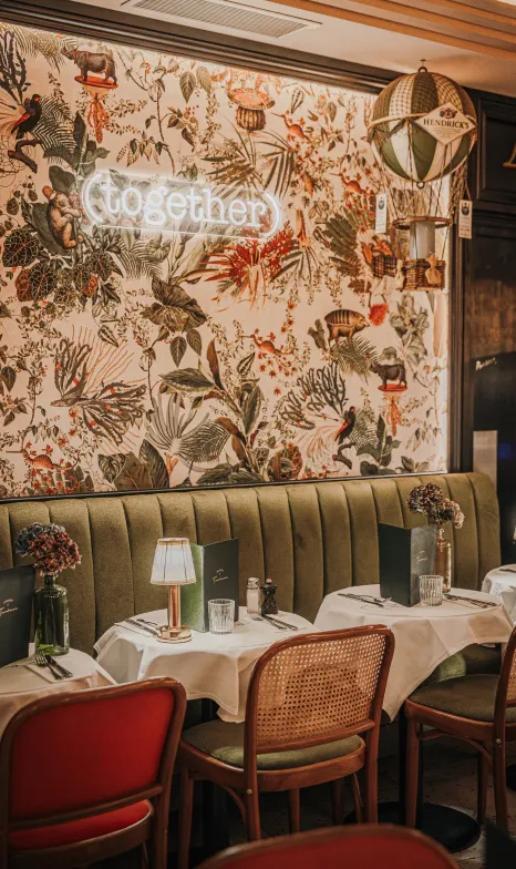 Cozy restaurant interior with floral wallpaper, neon sign reading 'together,' green upholstered bench seating, and tables set with menus, flowers, and a small lamp.