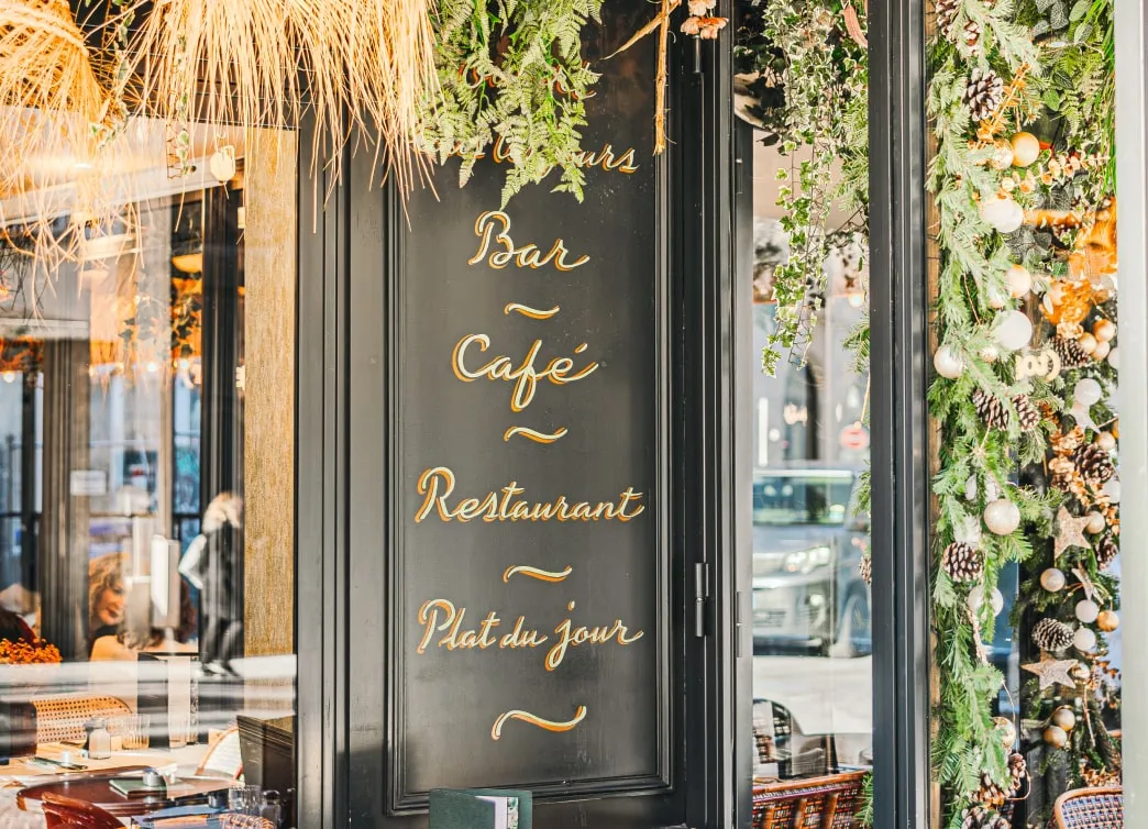 Black wooden sign on a restaurant window displaying the words 'Bar, Café, Restaurant, Plat du jour' with festive green garlands and pine cones decorating the window frame.