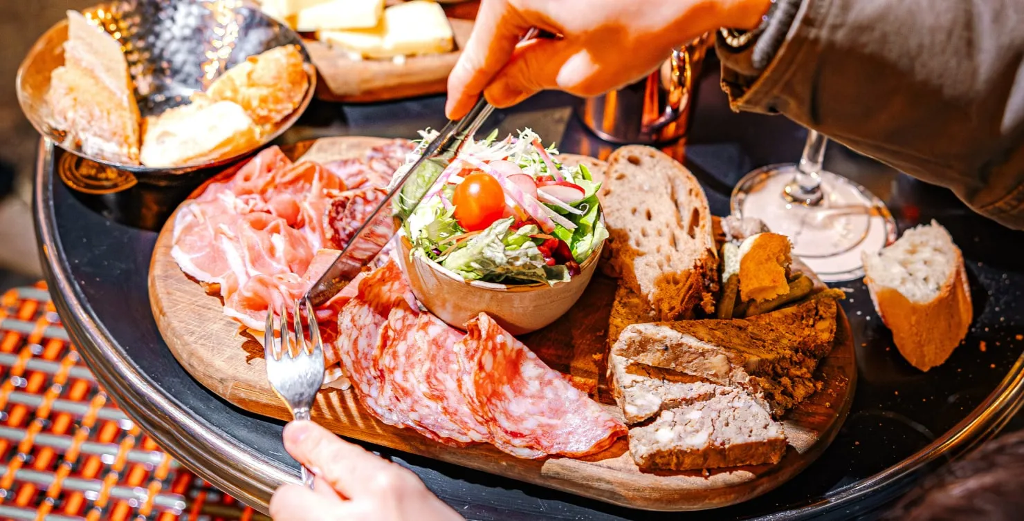 Charcuterie board with assorted cured meats, fresh salad with cherry tomato, rustic bread slices, pâté, and a bowl of bread pieces on a black table.