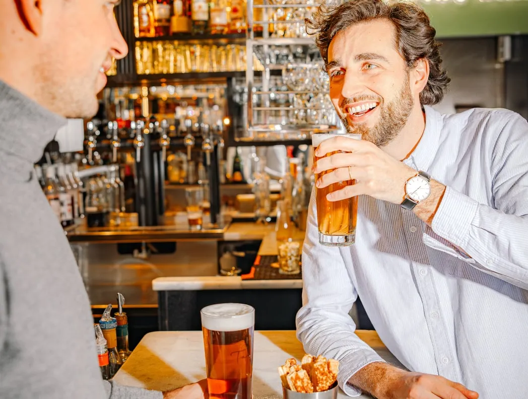 Two men smiling and enjoying beers at a bar counter with snacks in a metal cup.