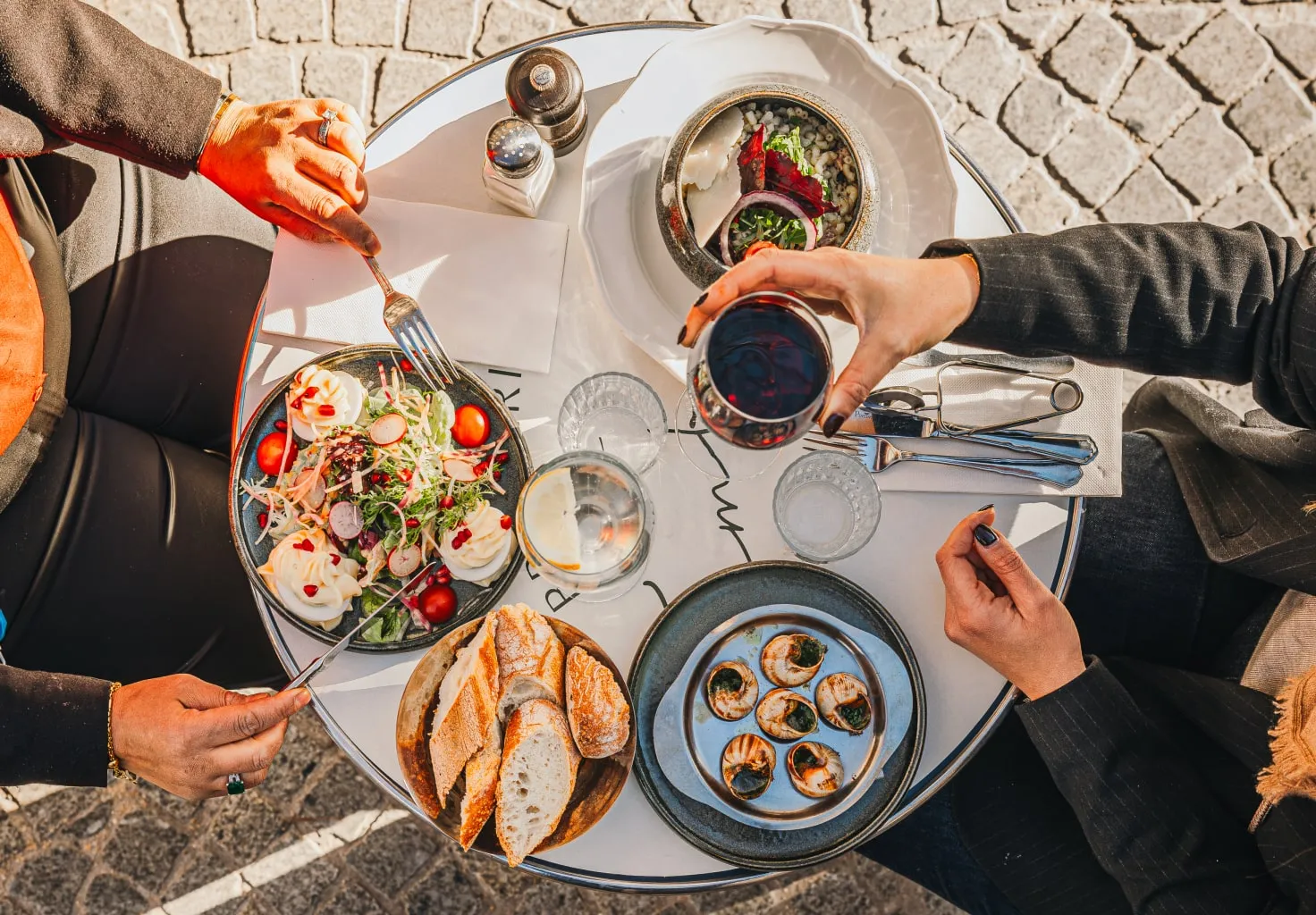 Overhead view of two people dining at a round table with salad, escargot, bread, and a glass of red wine.