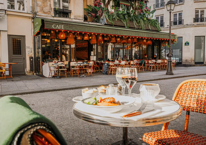 Outdoor table at a street cafe with a plate of food, a cup of coffee, and two glasses of water, with the cafe and empty chairs in the background.