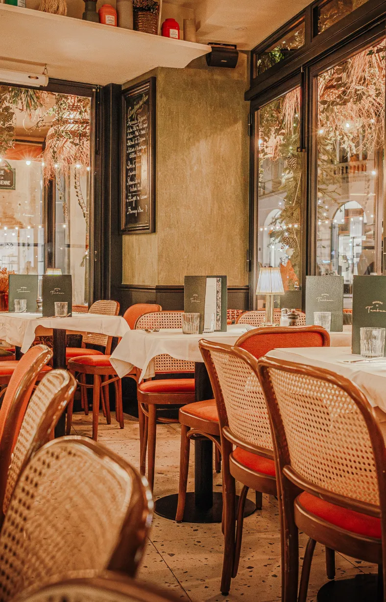 Cozy restaurant interior with wooden chairs featuring woven backs, white tablecloths, table lamps, menus, and large windows decorated with hanging plants and string lights.