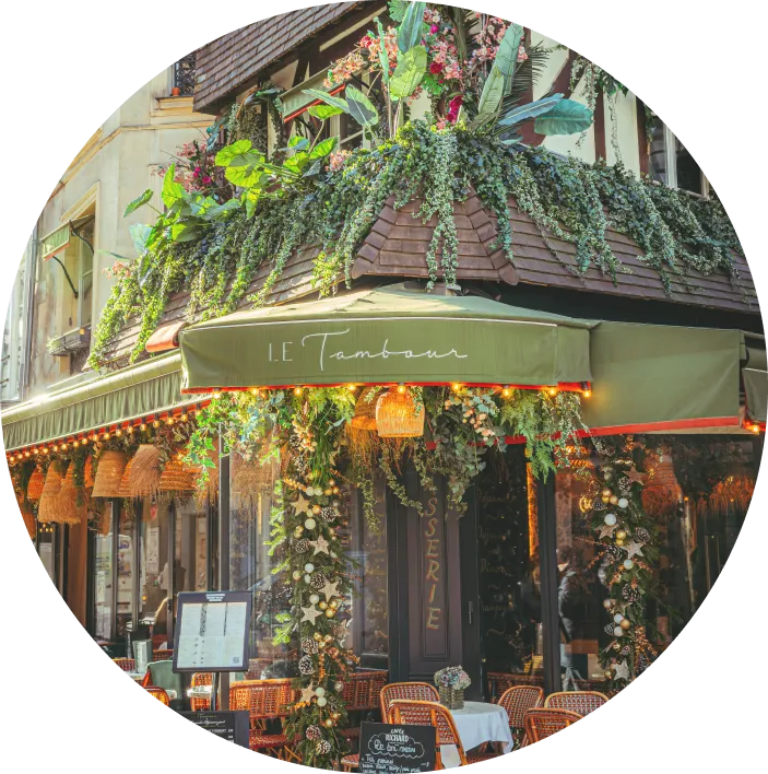 Outdoor corner of Le Tambour café with green awnings, hanging wicker lamps, plants, and tables with chairs set for dining.