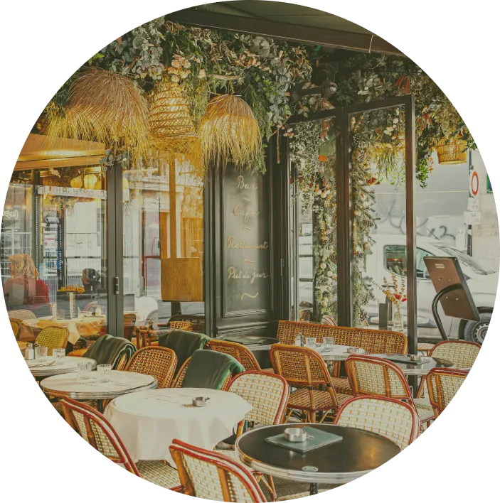 Cozy outdoor café seating area with rattan chairs, white tablecloths, hanging wicker lamps, and lush green foliage decor.