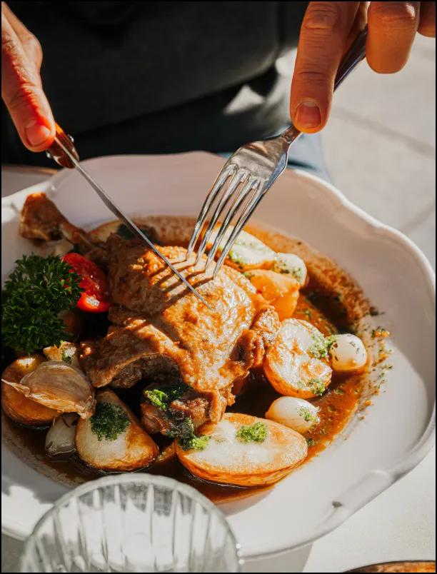 Person cutting into a cooked duck leg served with roasted potatoes, pearl onions, cherry tomatoes, and parsley garnish on a white plate.
