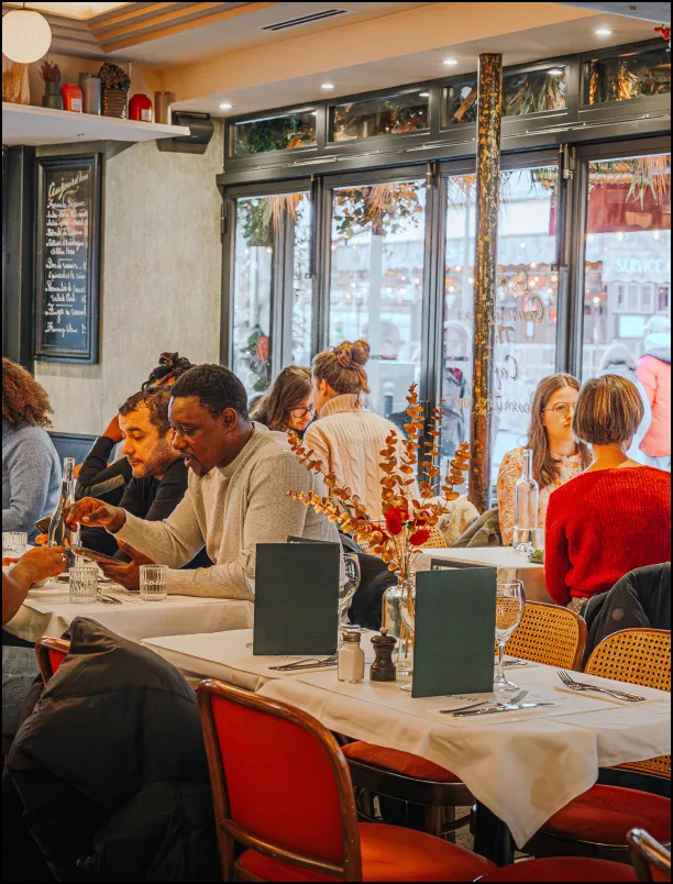 People sitting and dining at tables inside a cozy restaurant with large windows and warm lighting.