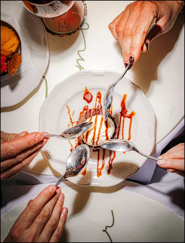 Four people holding spoons around a dessert plate with ice cream and caramel drizzle.