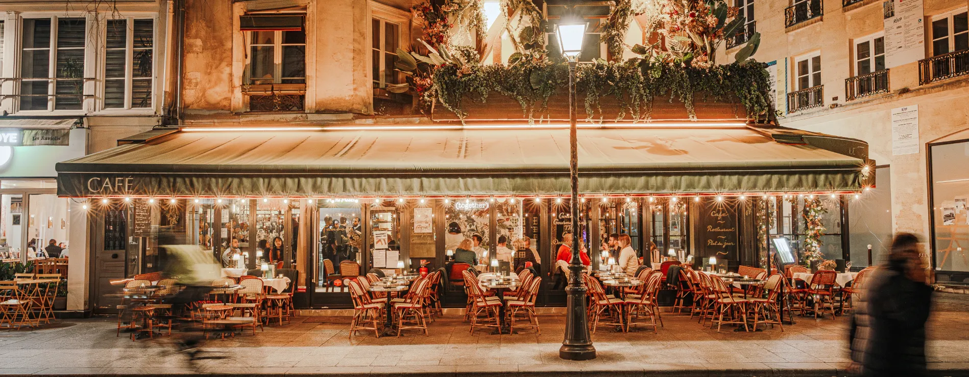 Outdoor seating area of a warmly lit café at night with patrons inside and blurred figures passing by.