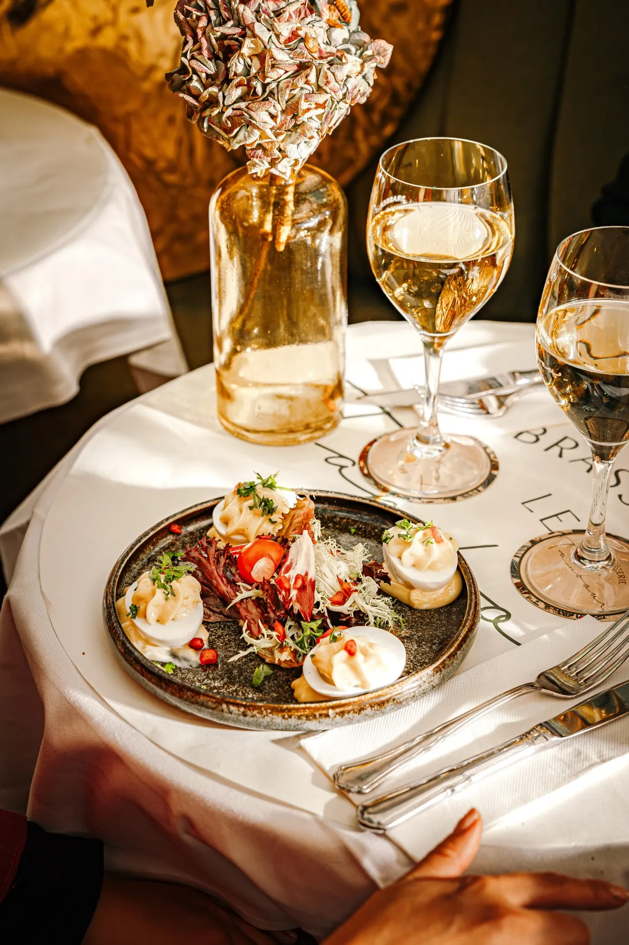 Plate with deviled eggs garnished with herbs and salad greens on a white tablecloth next to two wine glasses and a glass vase with dried flowers.