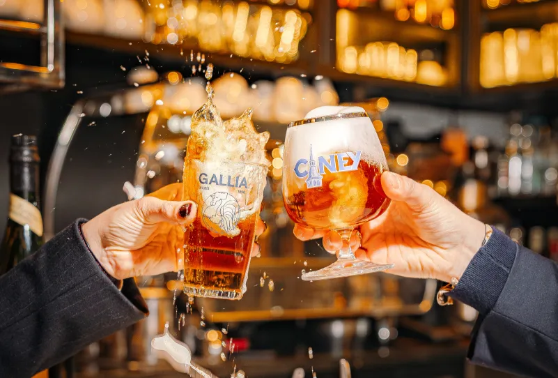 Two hands holding beer glasses clinking together with beer splashing in a lively bar setting.