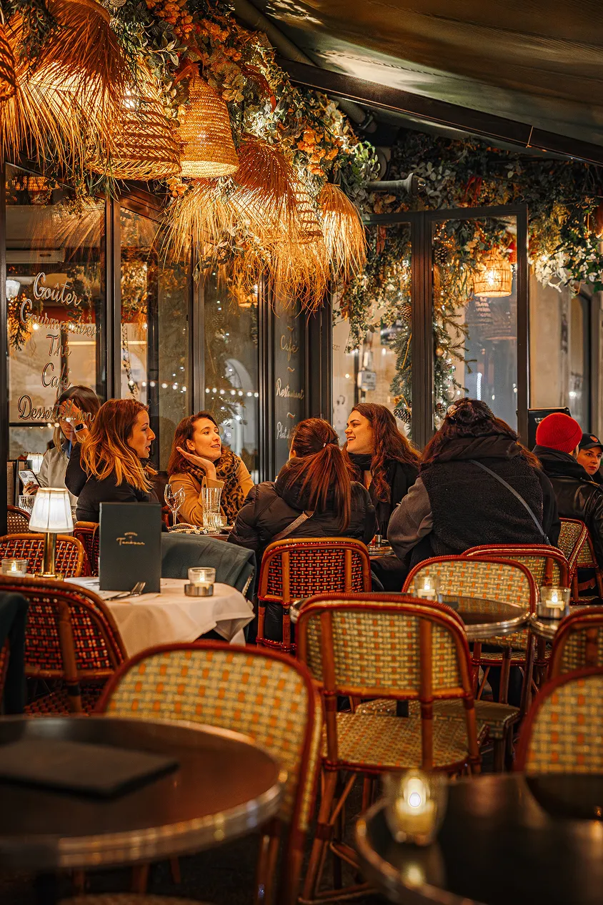 Group of people sitting and talking at an outdoor café decorated with hanging wicker lamps and plants at night.