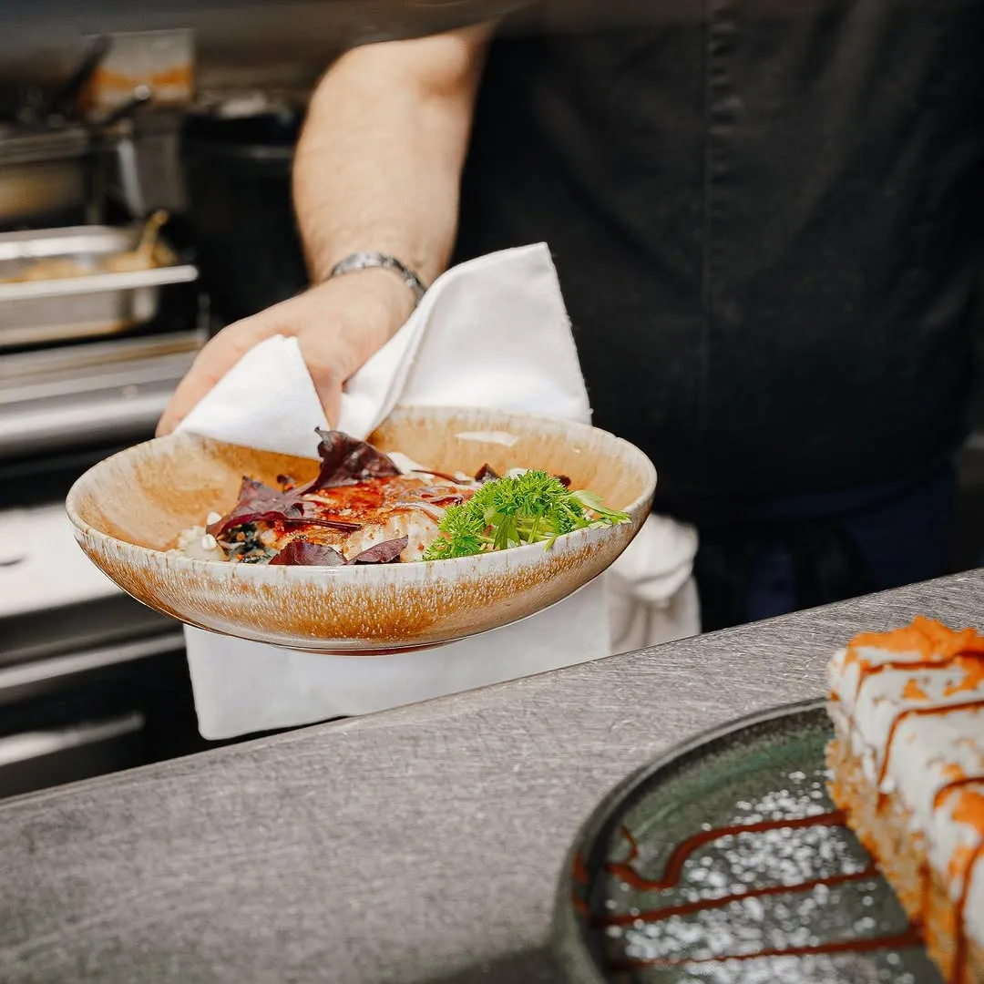 Person holding a beige bowl with a garnished dish, featuring greens and sauce, in a kitchen setting.
