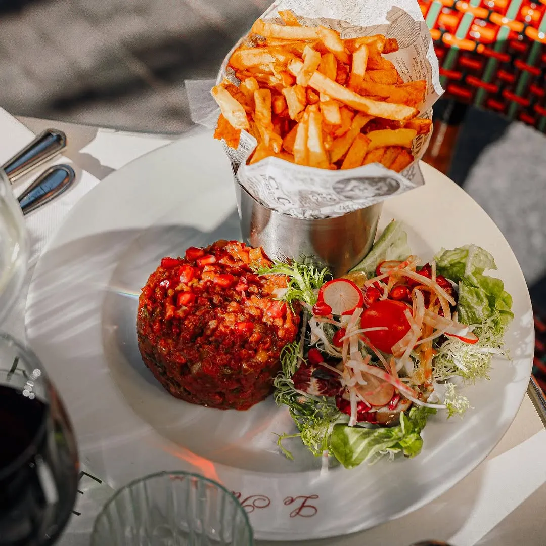 Plate with a serving of steak tartare garnished with pomegranate seeds, a mixed green salad with radishes and cherry tomato, and a metal cup filled with French fries.