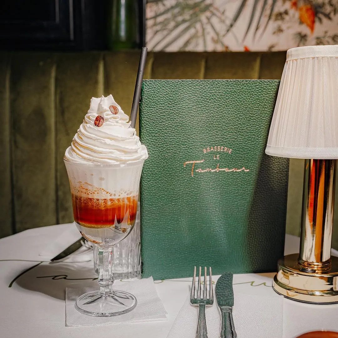 Glass of iced coffee topped with whipped cream and coffee beans on a white table with a green menu and a gold lamp nearby.