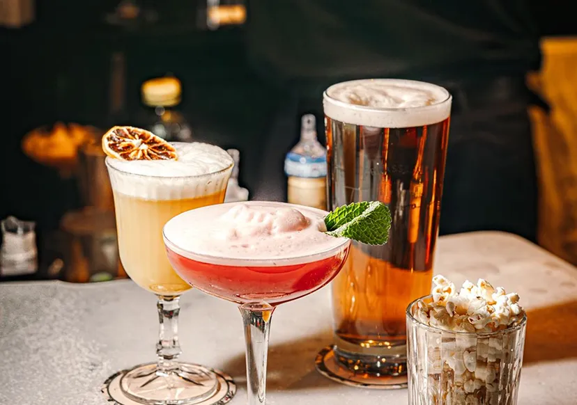 Four different drinks including a red cocktail with foam and mint, a yellow cocktail with foam and dried citrus, a glass of amber beer with foam, and a glass filled with popcorn.